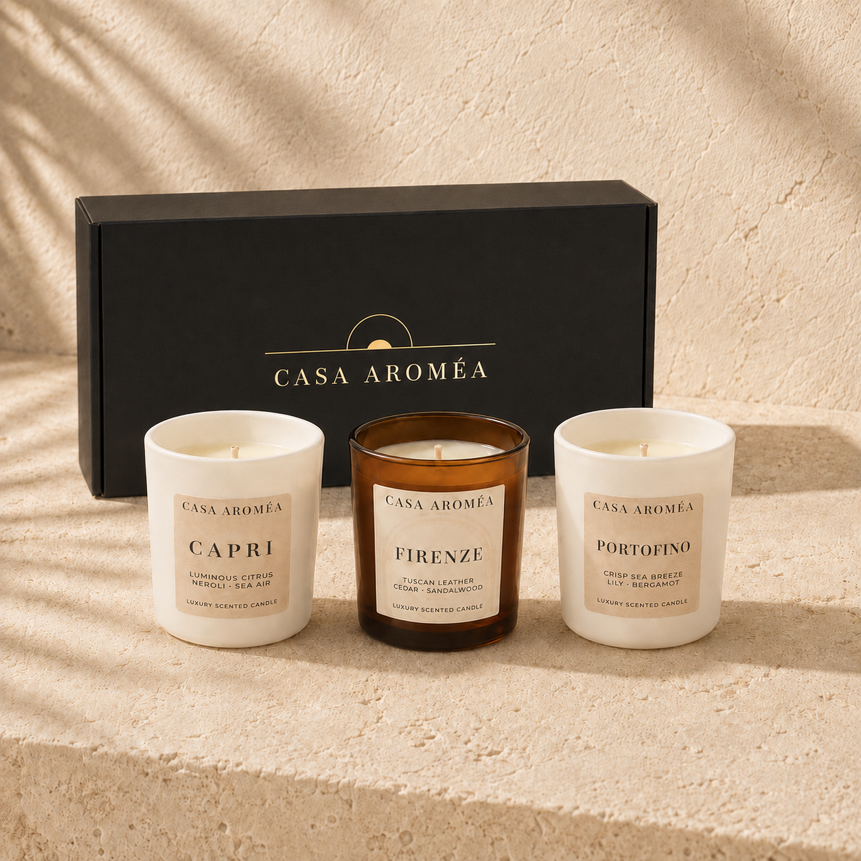 Three Casa Aroméa scented candles displayed in front of a black branded gift box on a textured beige surface. The left candle is white with a cream label reading Capri, the center candle is amber glass with a cream label reading Firenze, and the right candle is white with a cream label reading Portofino.