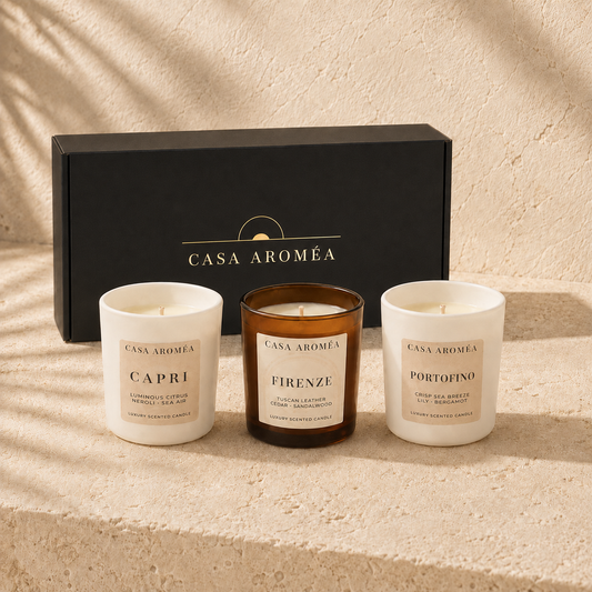 Three Casa Aroméa scented candles displayed in front of a black branded gift box on a textured beige surface. The left candle is white with a cream label reading Capri, the center candle is amber glass with a cream label reading Firenze, and the right candle is white with a cream label reading Portofino.